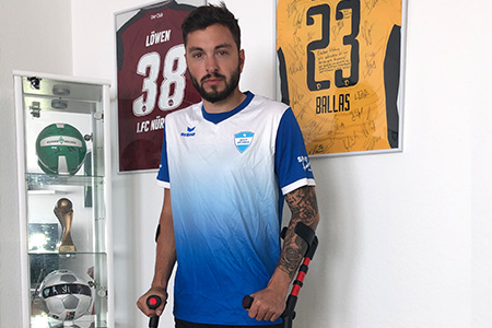 Beitragsbild Inklusions Challenge Gewinner: Einzelaufnahme von einem Amputierten Fußballer auf Krücken. Er steht vor einer Vitrine mit Bällen und Pokal und 2 an der Wand hängenden gerahmten Trikots